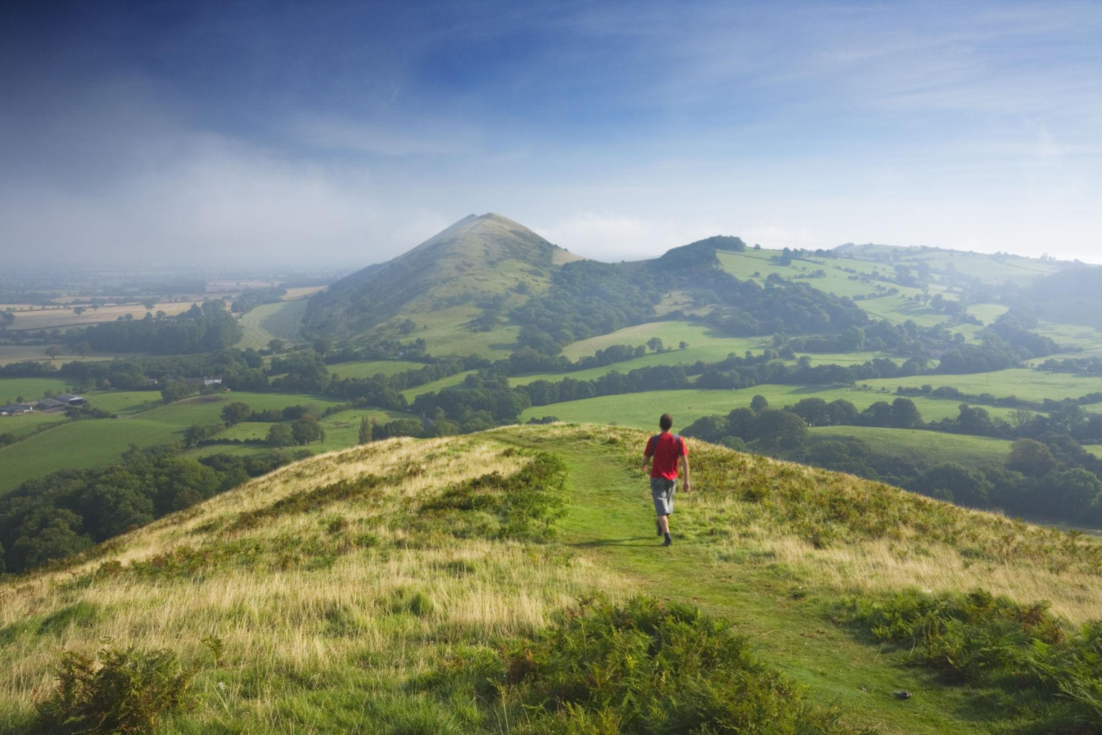 Walker following a  grassy ridge towards a pointed hill, overlooking green fields and wooded valleys.