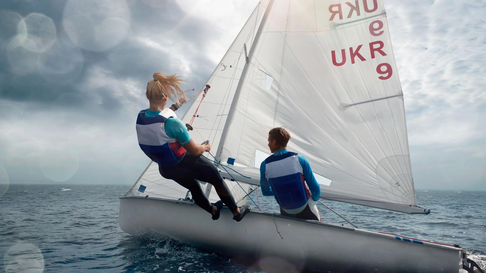 Two individuals in a sailboat on the water, with the sail displaying "UKR 9".