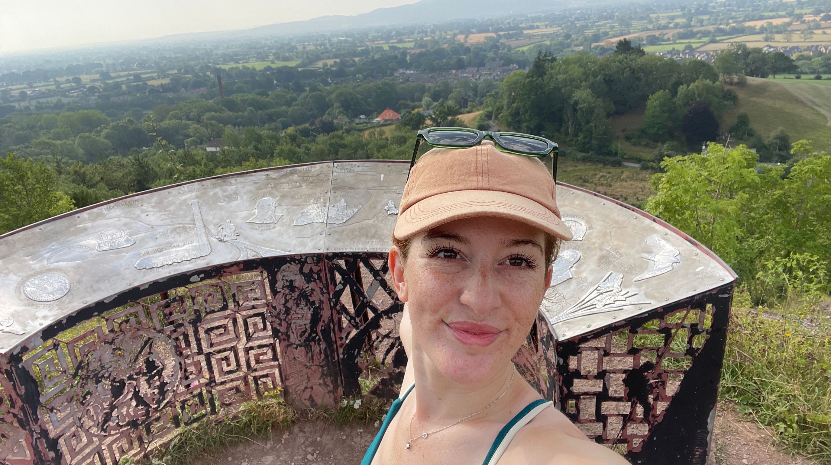 A person wearing a tan baseball cap and sunglasses takes a selfie with a scenic view of rolling hills and trees in the background. They are standing near a circular, textured structure.