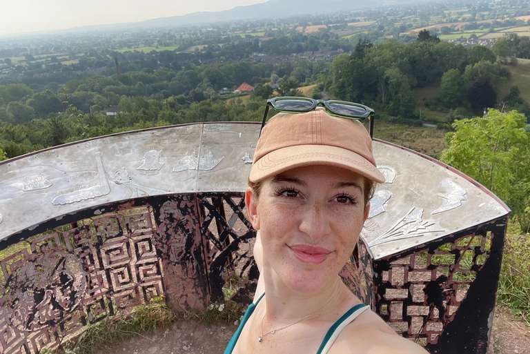 A person wearing a tan baseball cap and sunglasses takes a selfie with a scenic view of rolling hills and trees in the background. They are standing near a circular, textured structure.