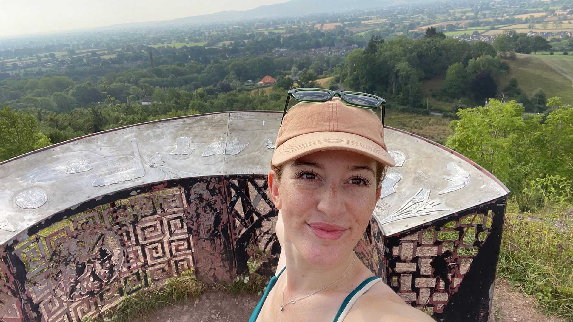 A person wearing a tan baseball cap and sunglasses takes a selfie with a scenic view of rolling hills and trees in the background. They are standing near a circular, textured structure.