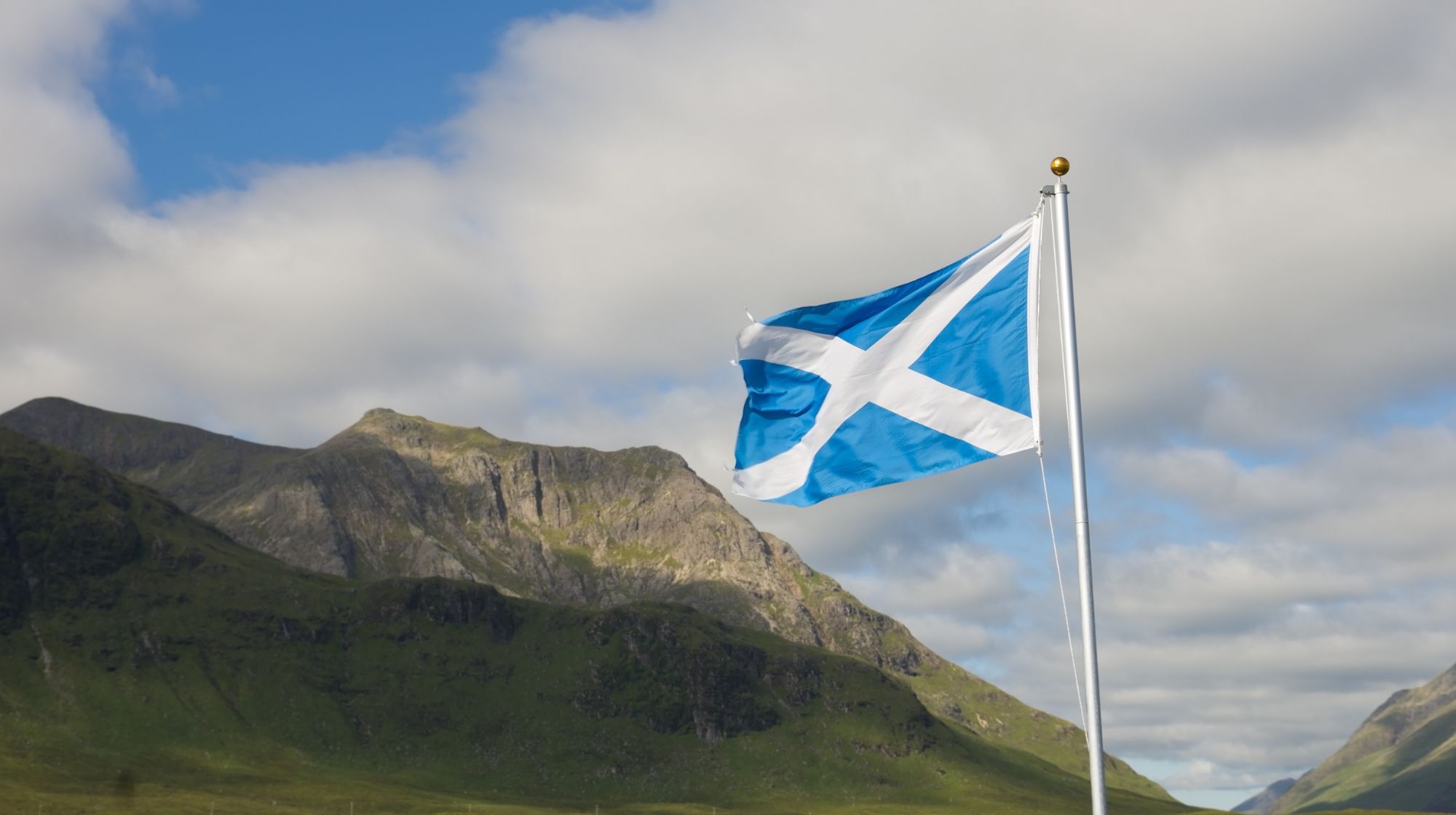 The Scottish flag, a blue background with a white saltire, flies in the foreground against a backdrop of green hills and mountains under a cloudy sky.