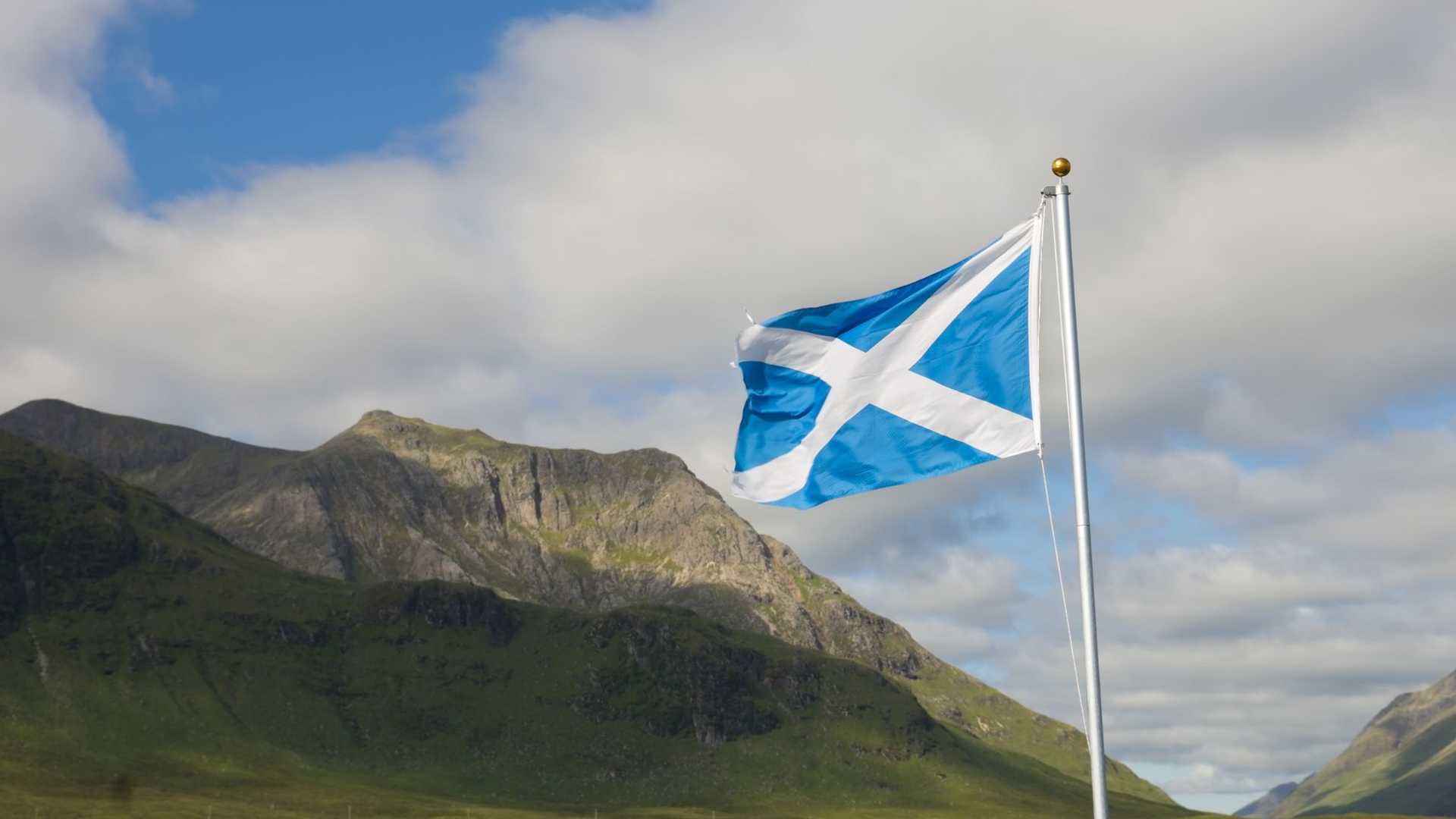 The Scottish flag, a blue background with a white saltire, flies in the foreground against a backdrop of green hills and mountains under a cloudy sky.