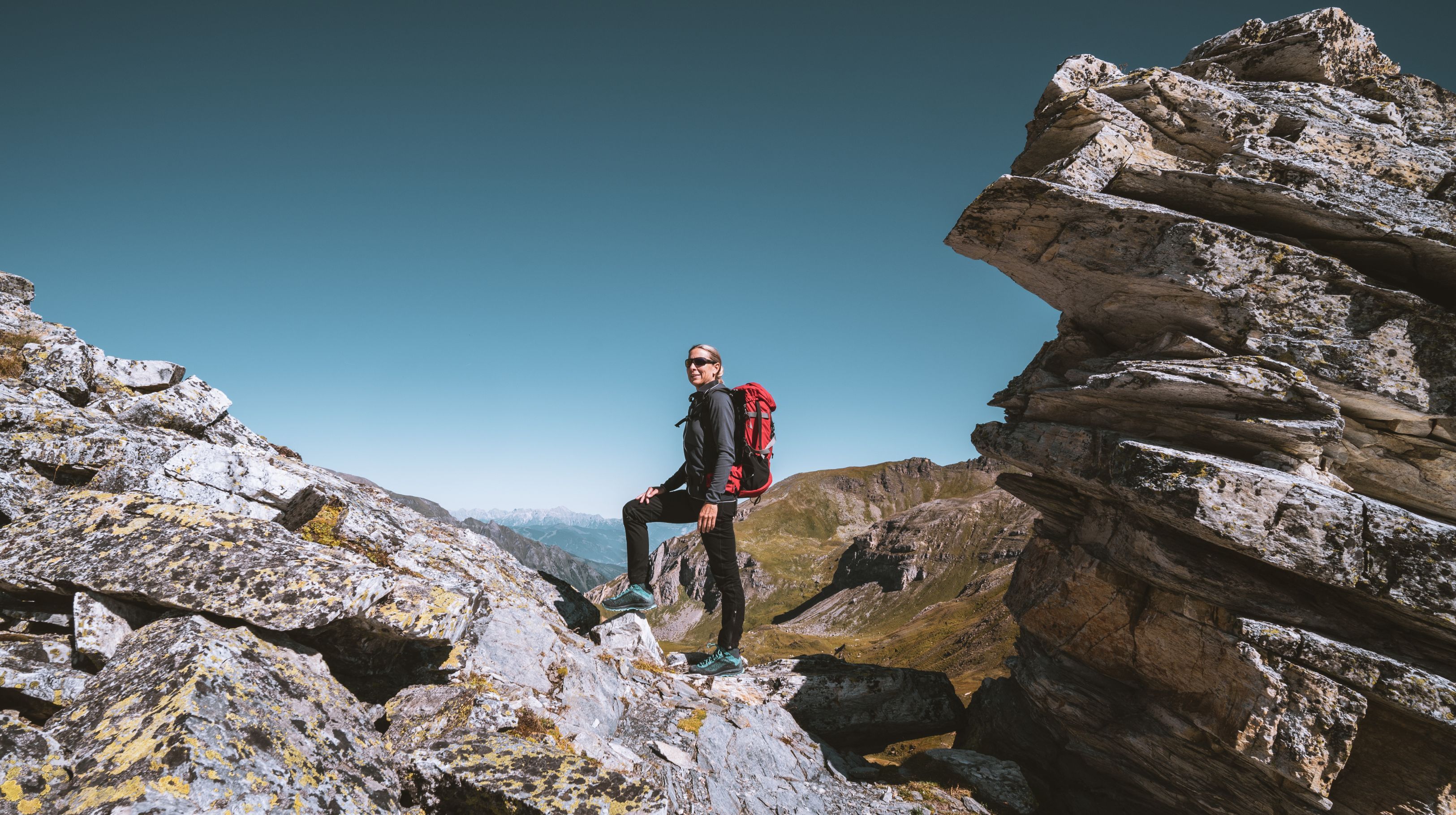 A person wearing a backpack stands on a rocky mountain path with a clear blue sky above.