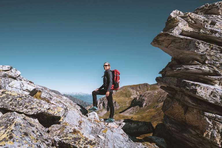A person wearing a backpack stands on a rocky mountain path with a clear blue sky above.