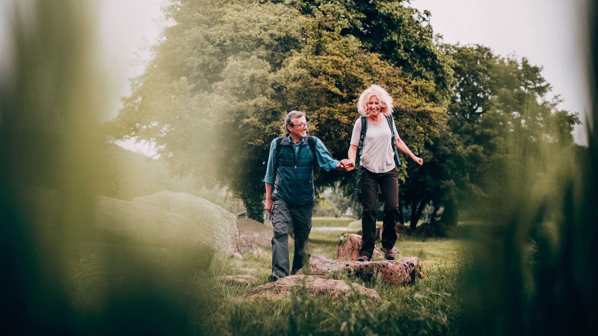 A couple holding hands while hiking on a rocky path in a park with trees in the background.