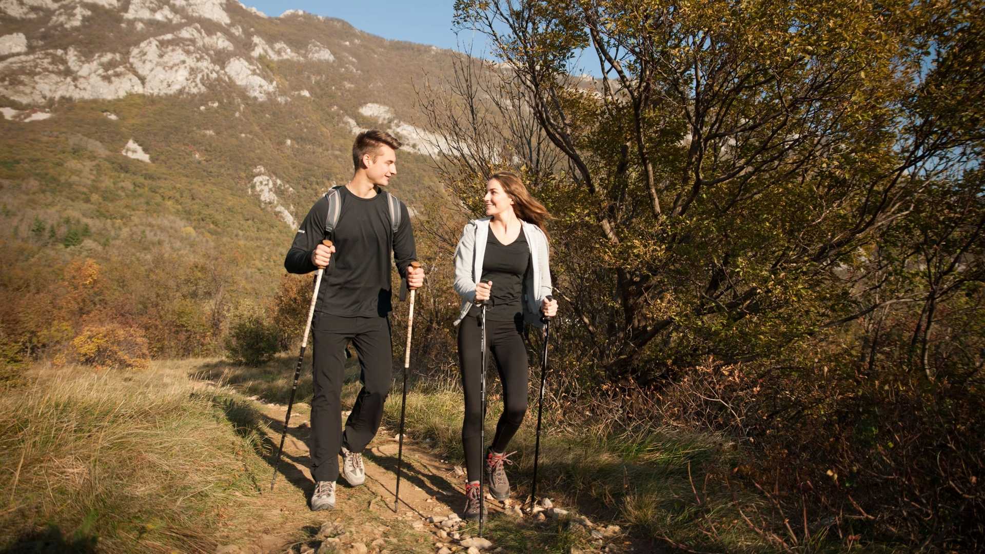 Two hikers with trekking poles walk on a dirt path through a wooded area with mountains in the background.