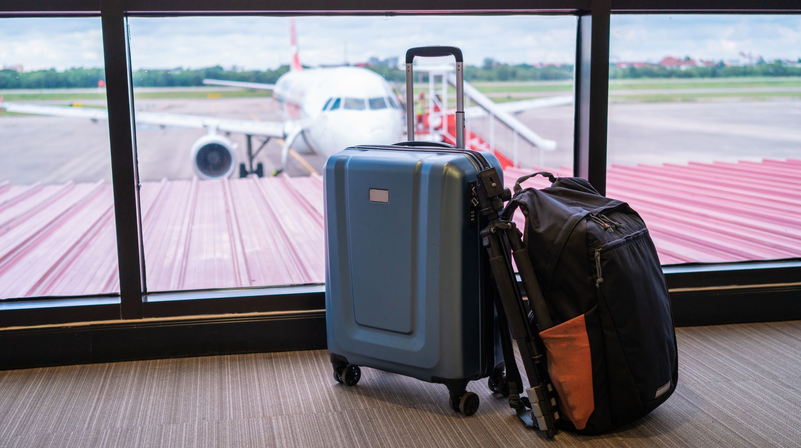 A blue rolling suitcase and a black backpack with an orange accent sit by a window overlooking an airplane on the tarmac.