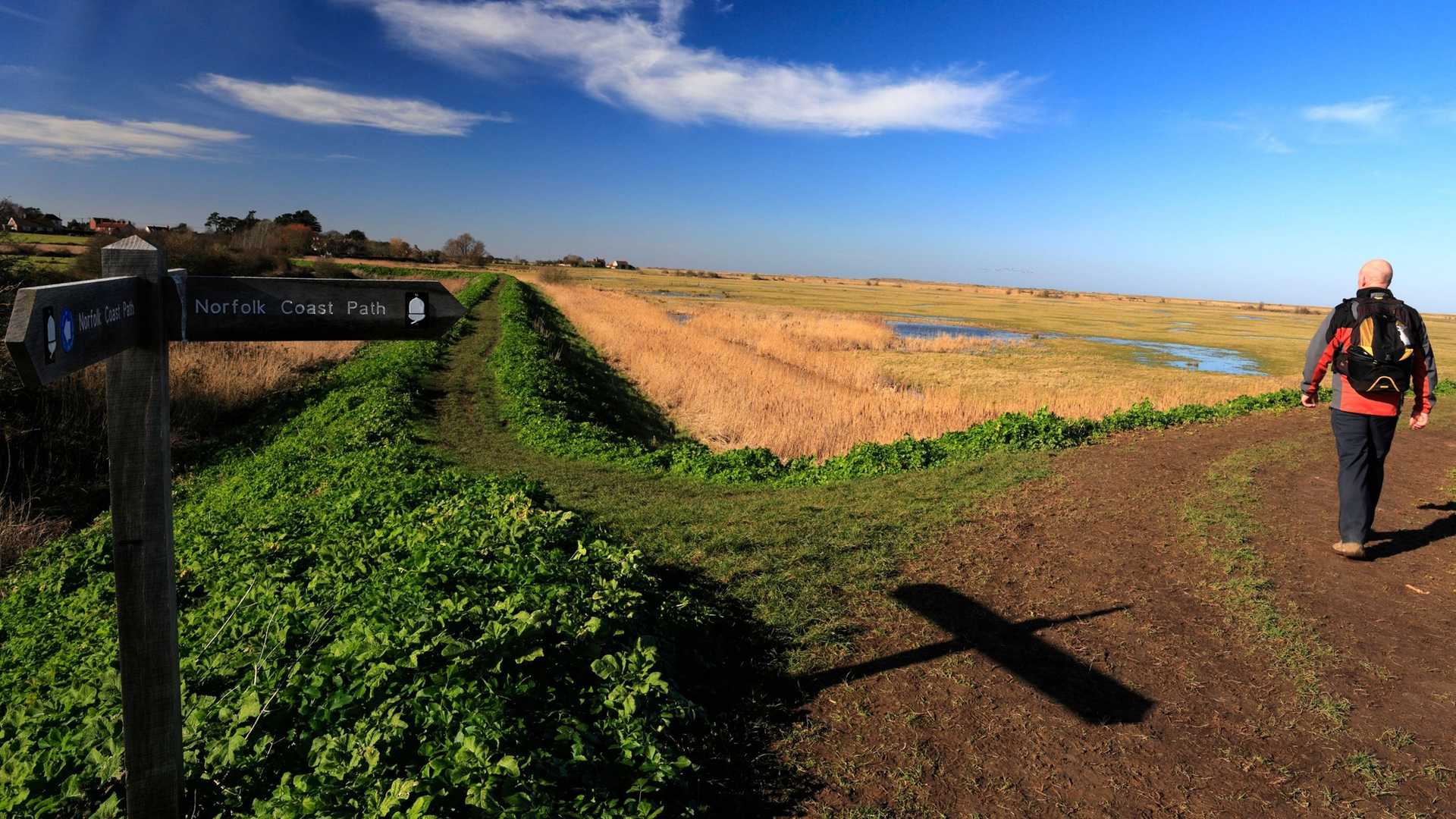A wooden signpost points towards the Norfolk Coast Path as a person walks away on a dirt path next to marshland under a blue sky with clouds.