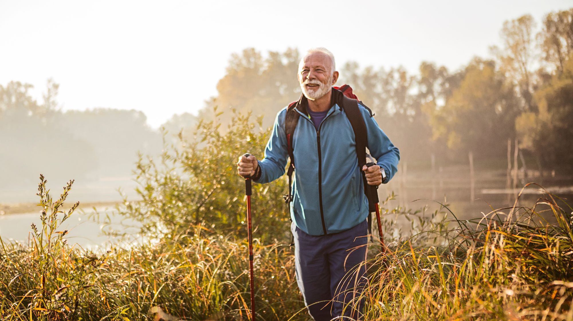 A man with a beard wearing a blue jacket and backpack hikes with trekking poles through tall grass near a body of water.