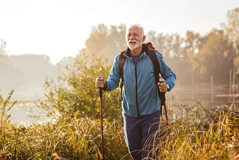 A man with a beard wearing a blue jacket and backpack hikes with trekking poles through tall grass near a body of water.