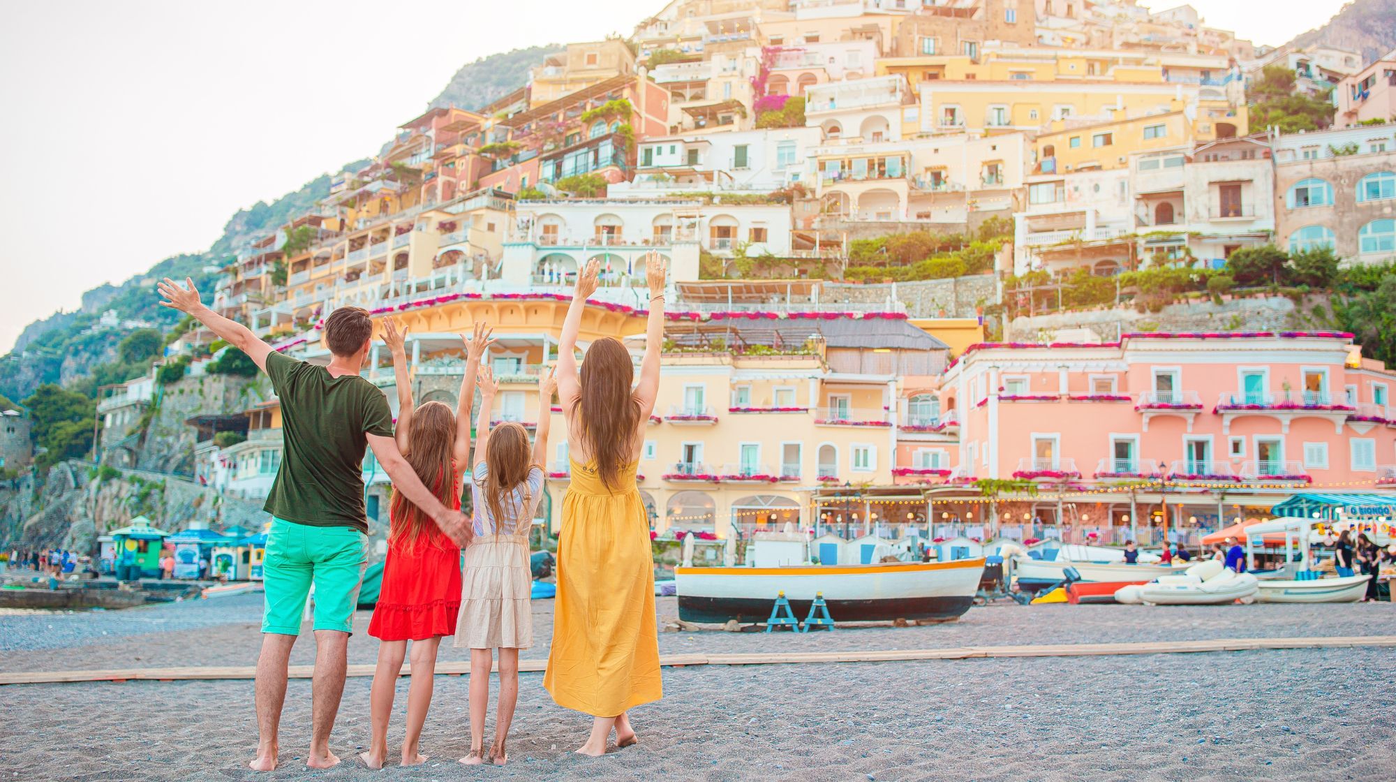 A family stands on a beach with their arms raised, facing a hillside town with colorful buildings and boats docked offshore.