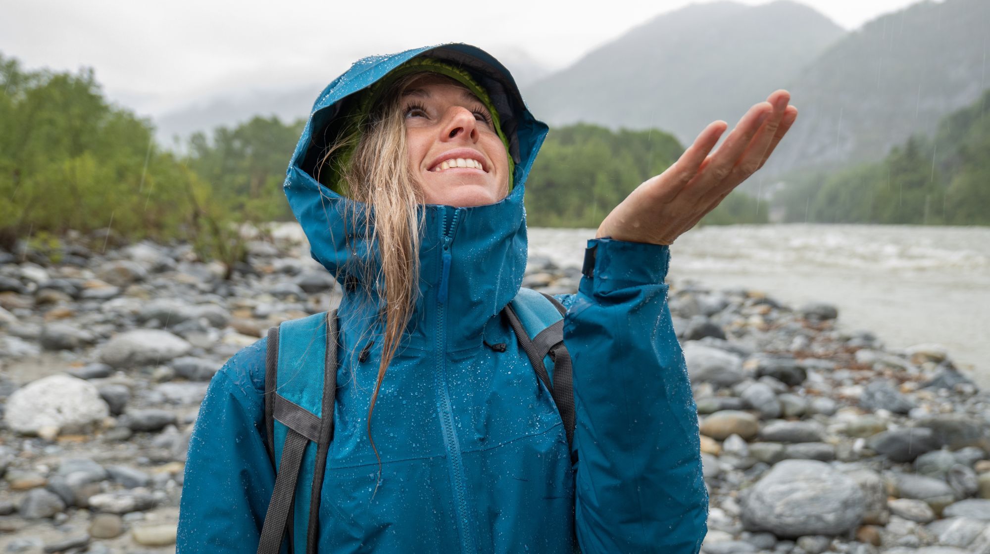 A person in a blue rain jacket smiles and holds out their hand to feel the rain on a rocky riverbank