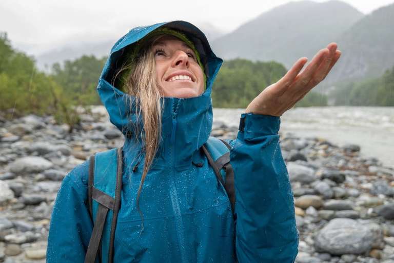 A person in a blue rain jacket smiles and holds out their hand to feel the rain on a rocky riverbank