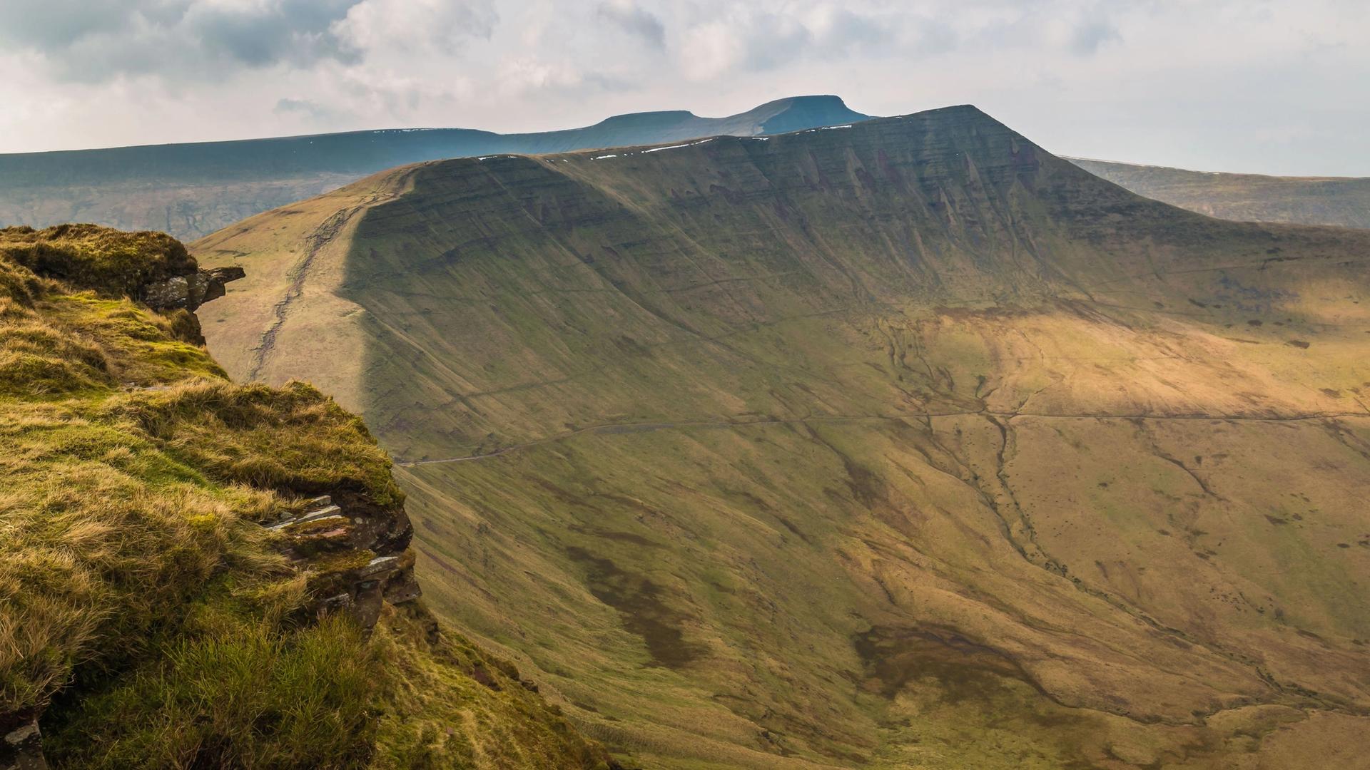 A wide shot of rolling hills and mountains under a cloudy sky. A grassy cliff edge is in the foreground.