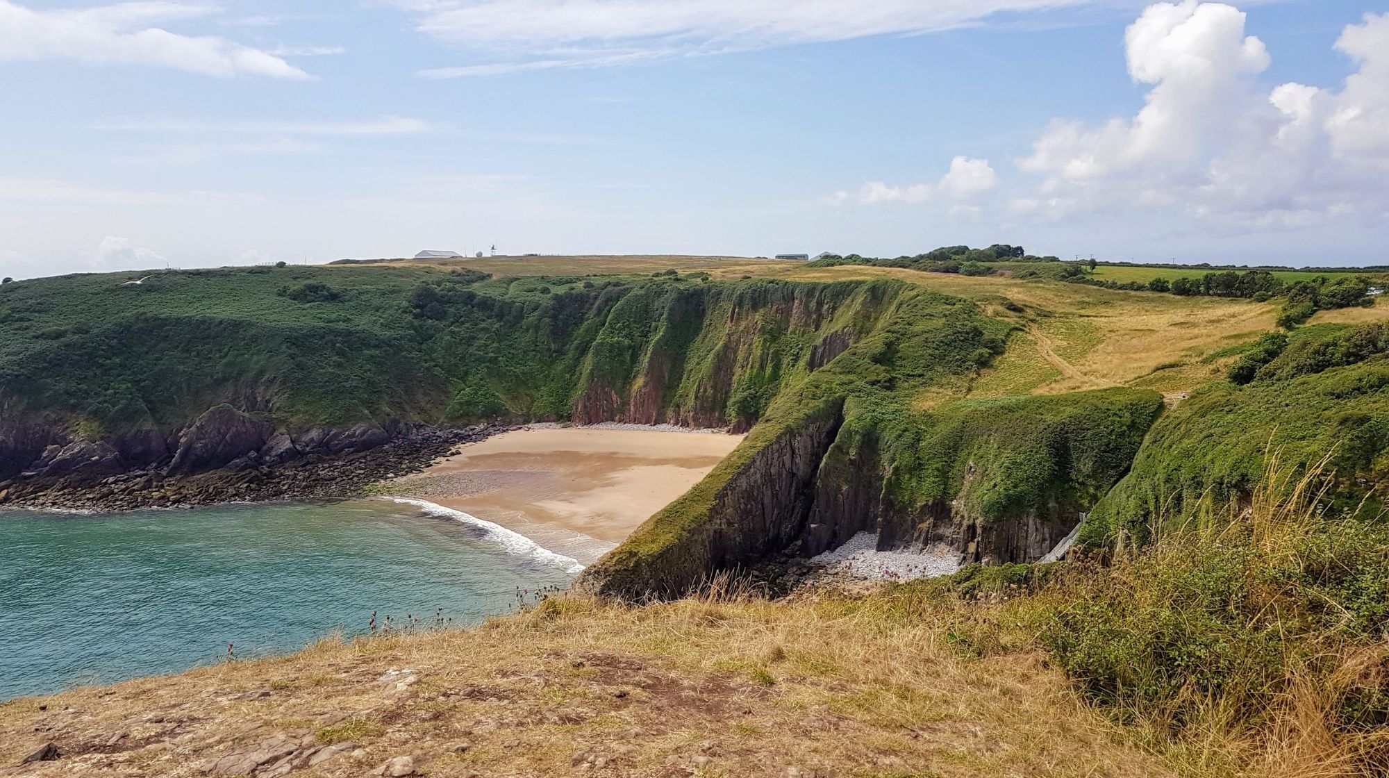 A view of a sandy cove surrounded by green, grassy cliffs under a partly cloudy sky.