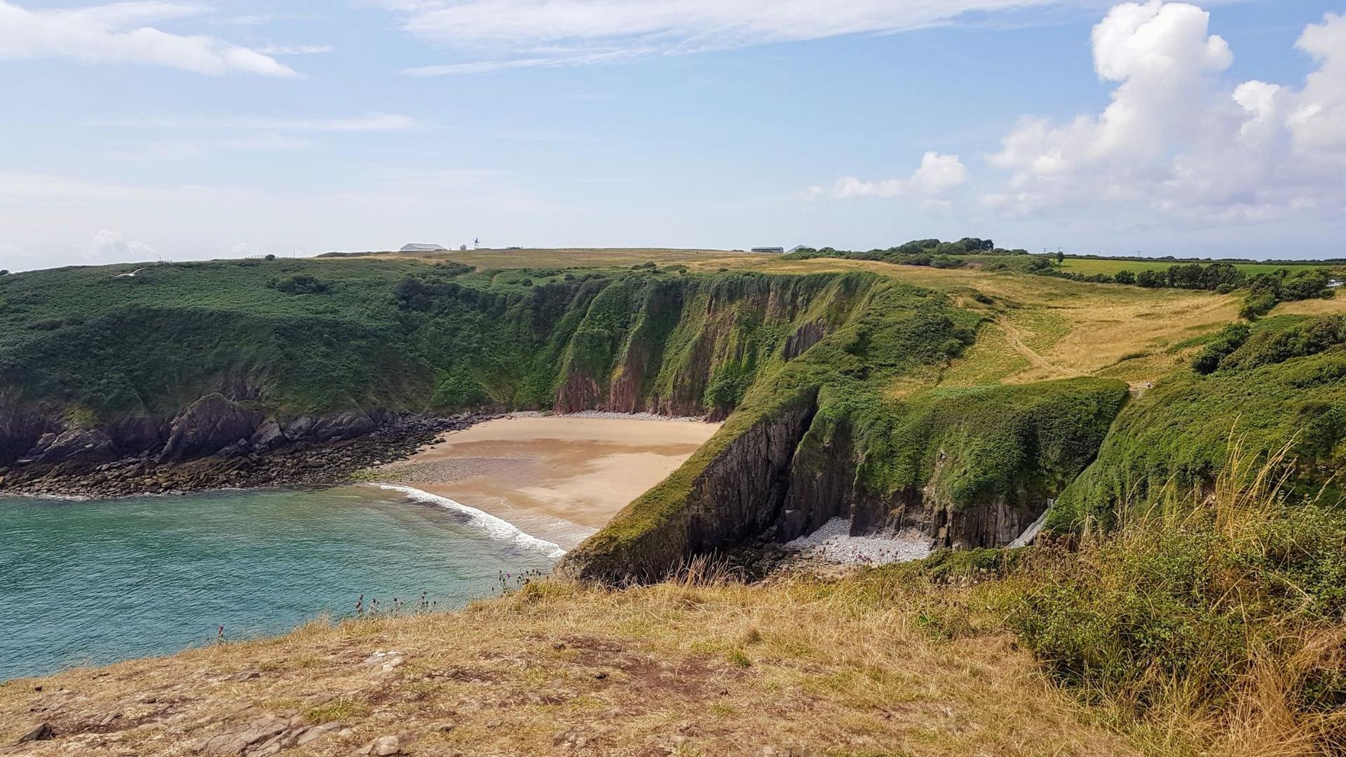 A view of a sandy cove surrounded by green, grassy cliffs under a partly cloudy sky.