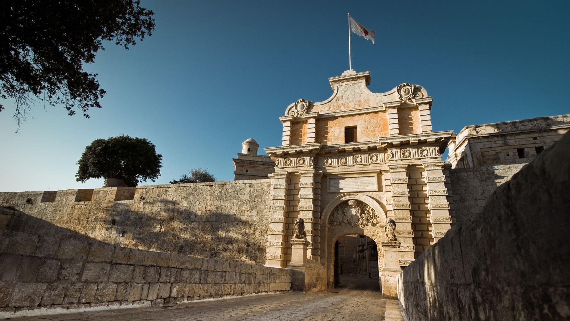 A grand stone gatehouse with a flag flying from the top under a clear blue sky..