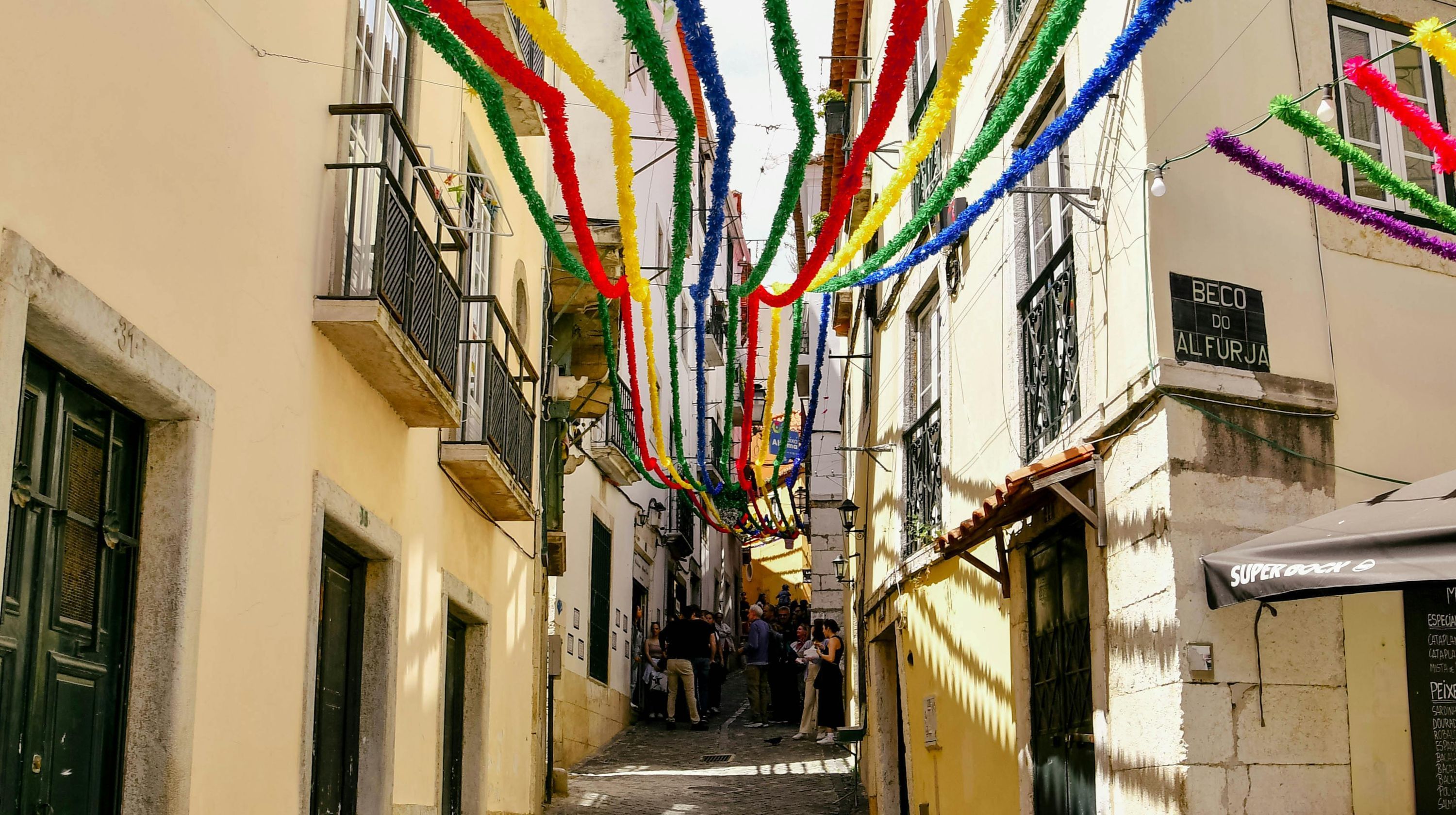 A narrow street decorated with colorful streamers strung between buildings. The buildings have balconies and windows, and there are people walking in the distance.