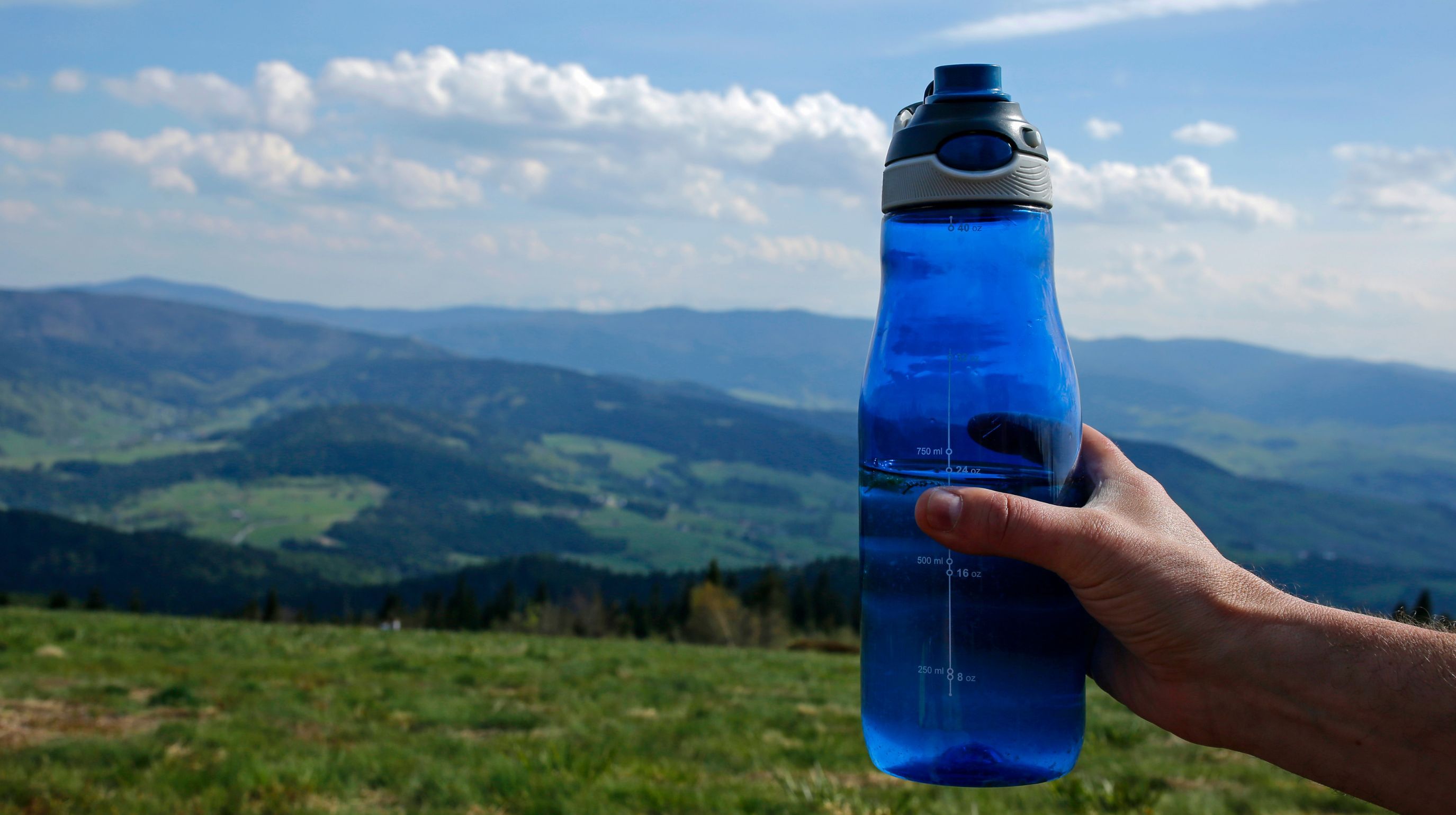 A hand holds a blue water bottle against a backdrop of rolling green hills and a cloudy sky.