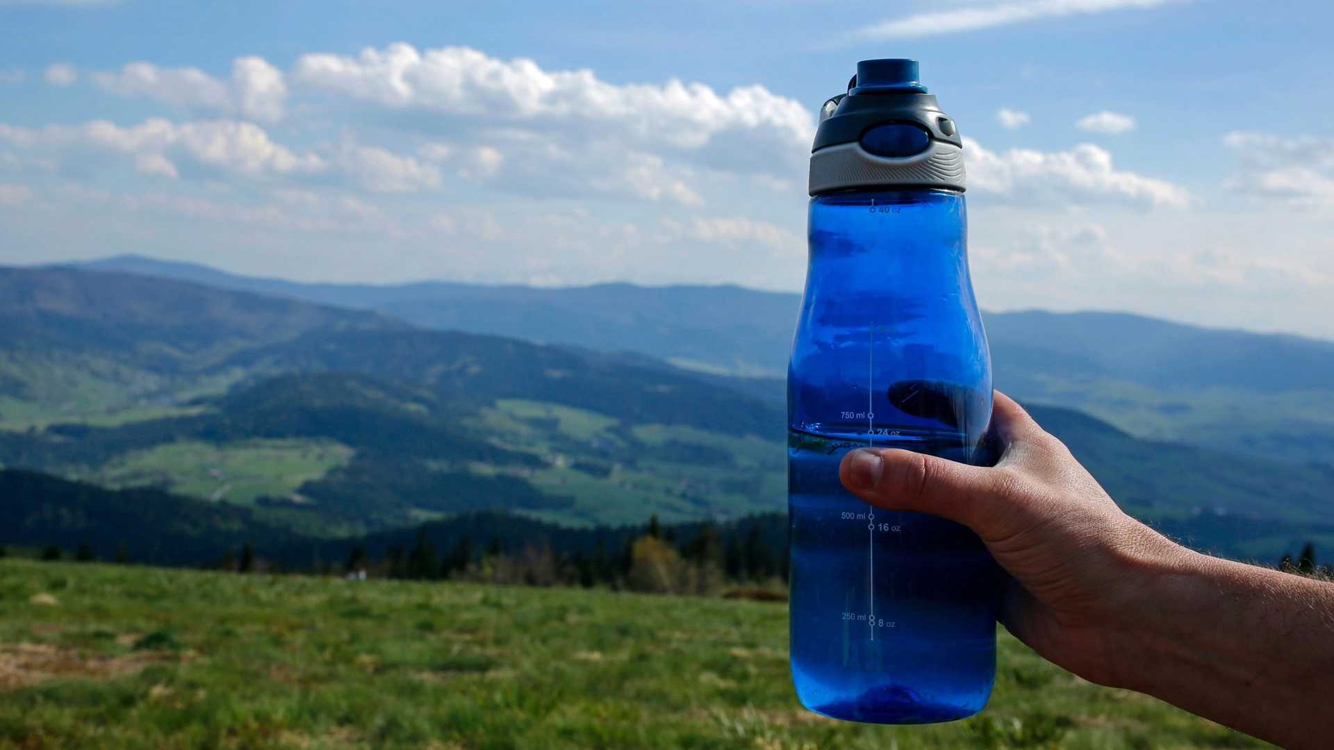 A hand holds a blue water bottle against a backdrop of rolling green hills and a cloudy sky.