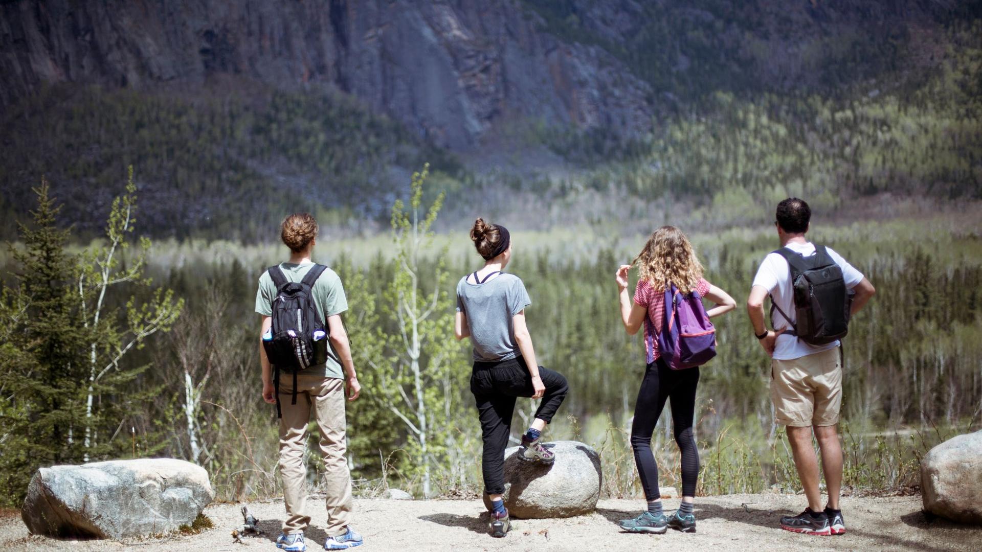 Four hikers stand on a rocky outcrop, looking out at a vast valley with a steep, dark mountain face in the distance