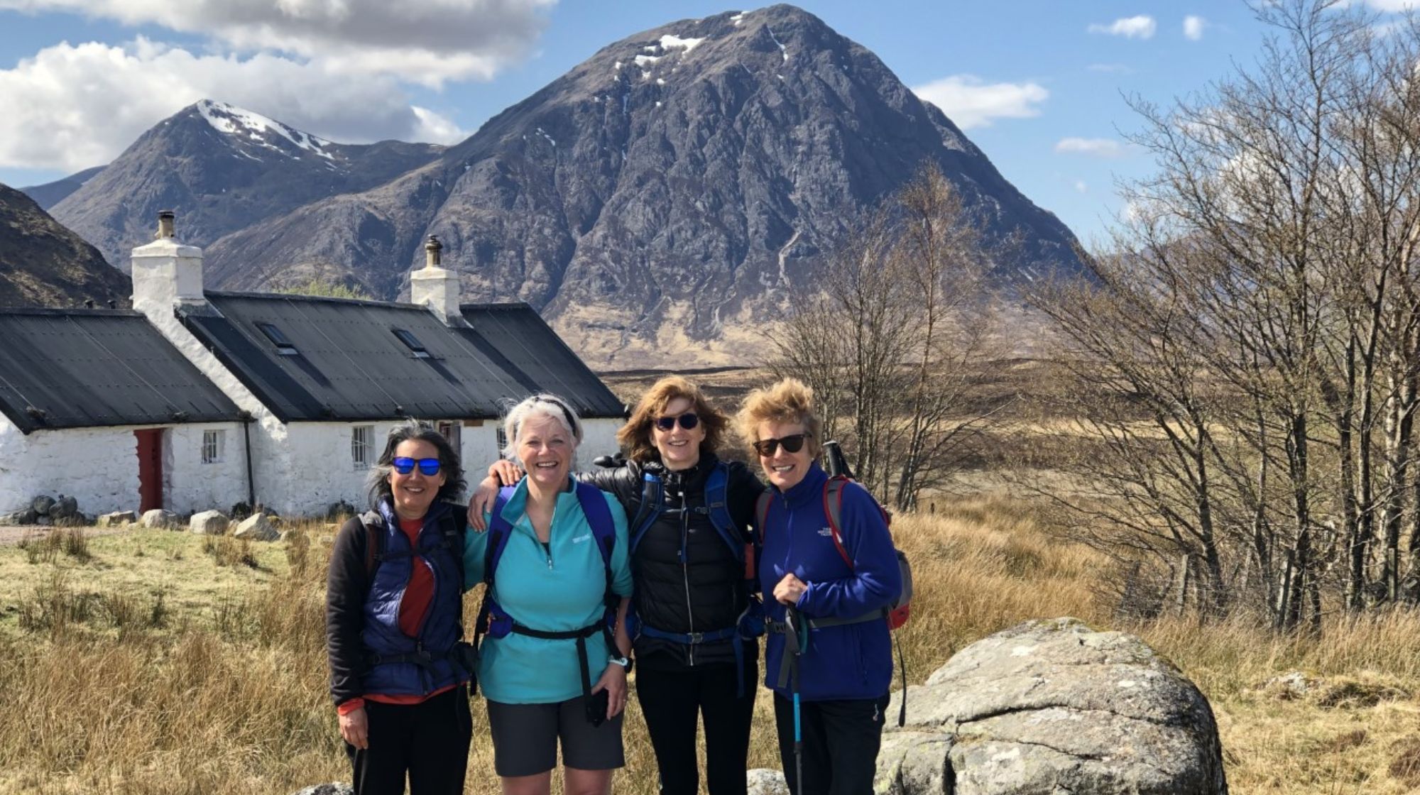 Four hikers stand in front of white cottages with dark roofs, a large mountain in the background, and bare trees to the right.