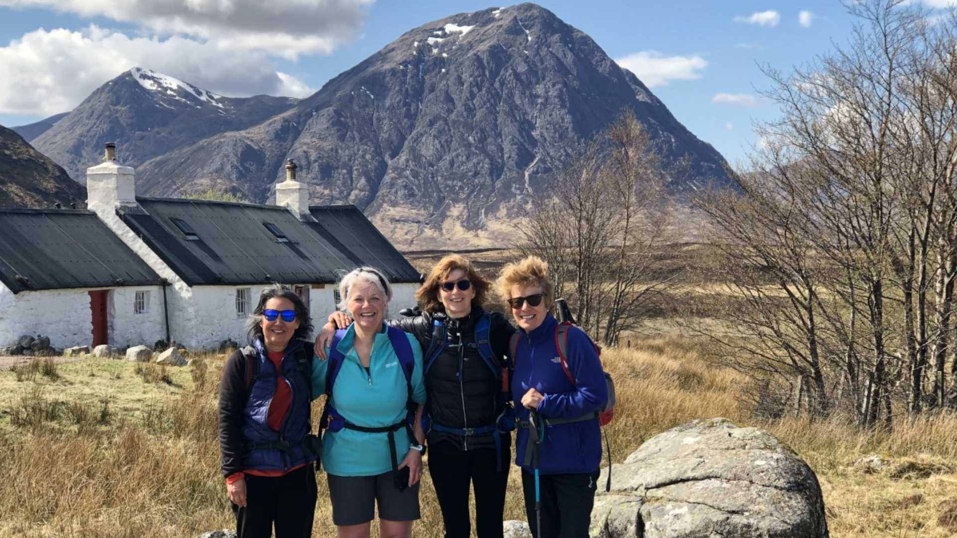 Four hikers stand in front of white cottages with dark roofs, a large mountain in the background, and bare trees to the right.