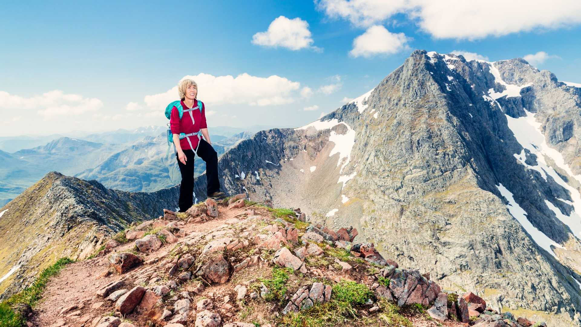 A hiker stands on a rocky mountain peak, looking out at a vast landscape of rolling mountains under a blue sky with scattered clouds.