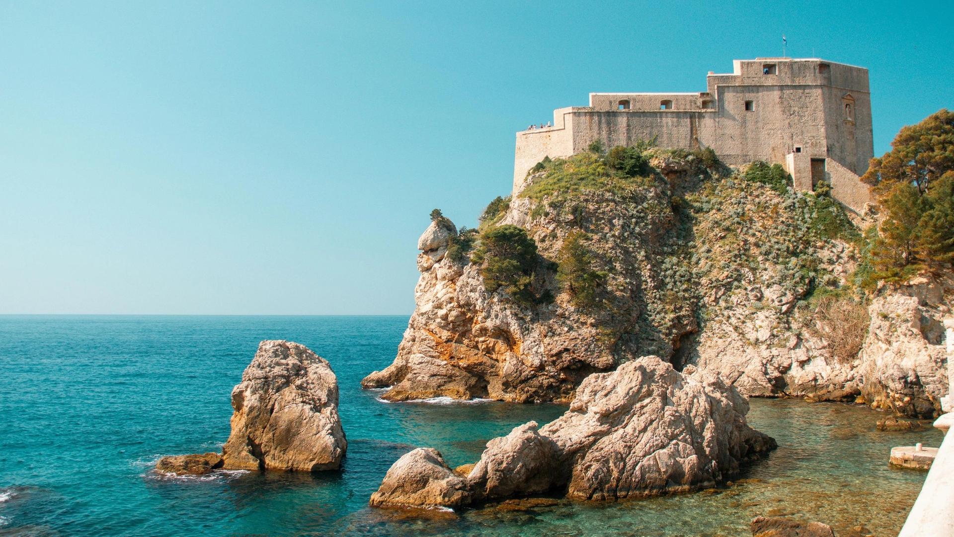 A stone fortress sits atop a rocky cliff overlooking the turquoise ocean. Several large rocks are visible in the water near the shore.
