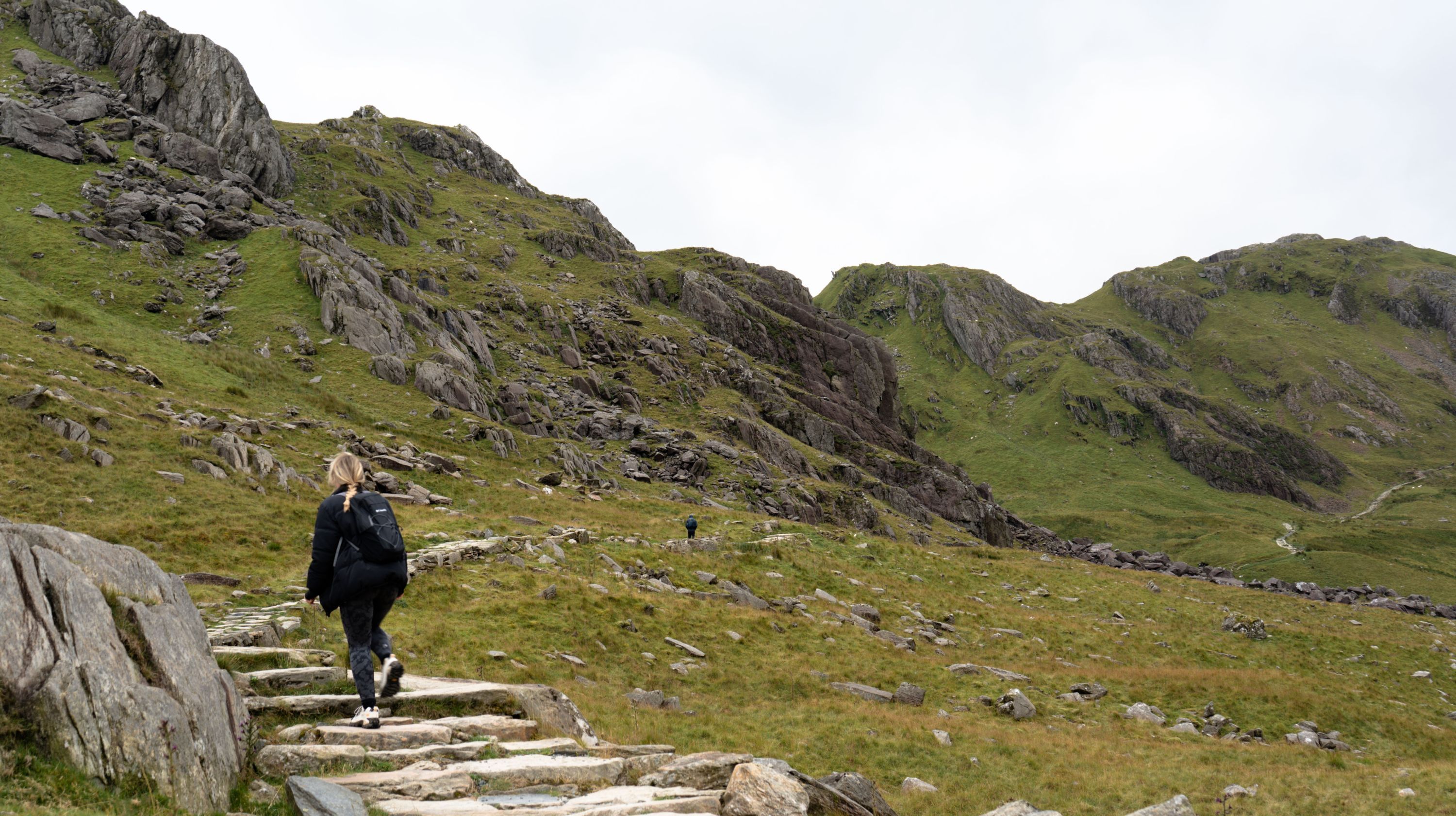 A person walks up a stone staircase on a grassy mountainside under a cloudy sky.