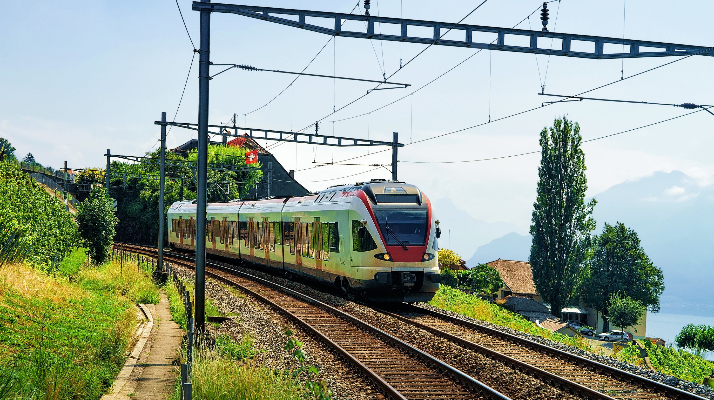 A modern train travels on tracks through a scenic landscape with lush green hills, trees, and distant mountains under a clear blue sky.