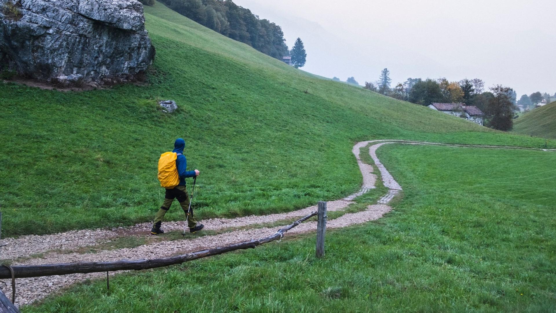 A hiker with a yellow backpack walks along a dirt path through a green, hilly landscape, using trekking poles for support