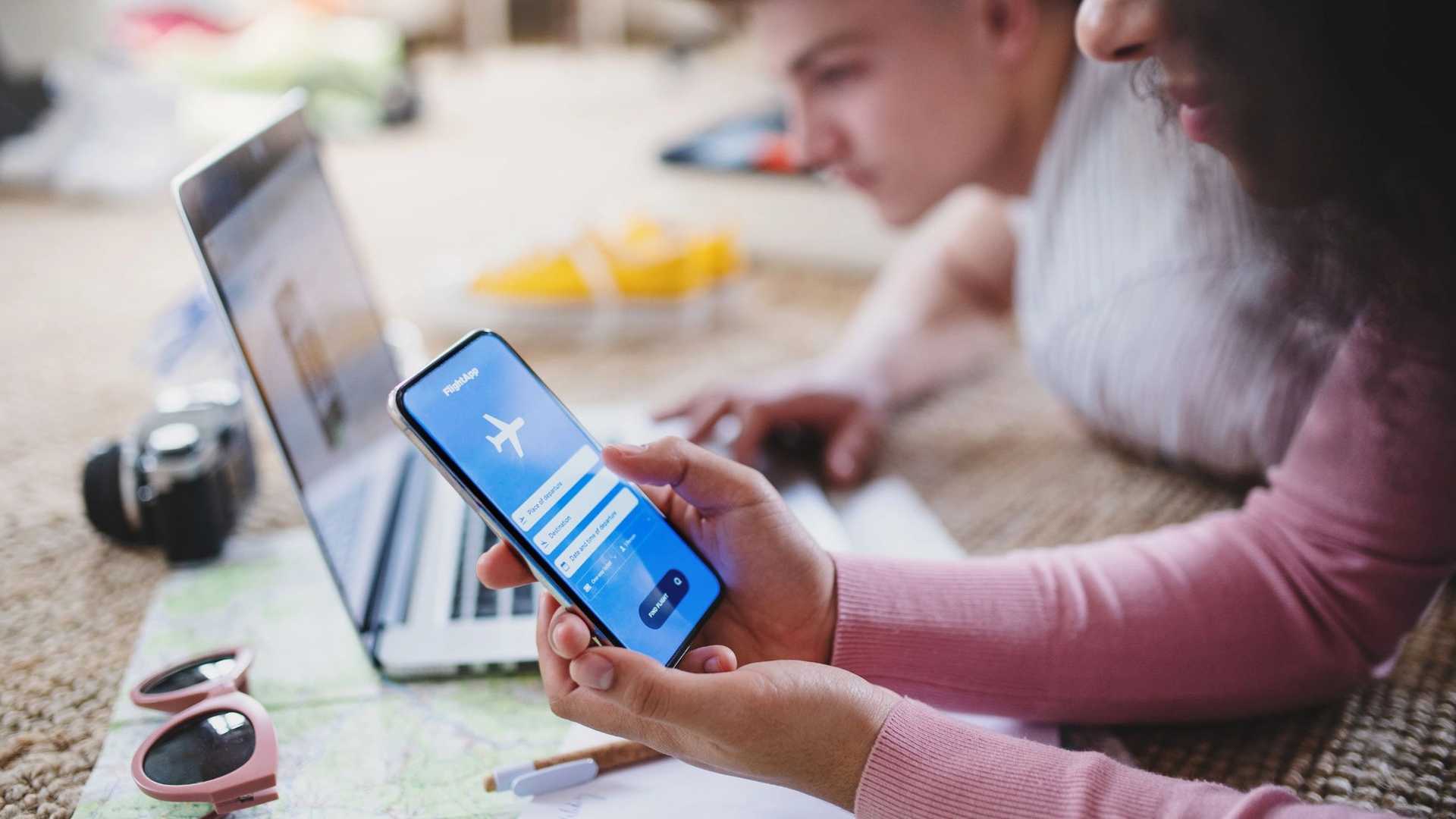 A person holds a smartphone displaying an airplane icon and booking fields, while a laptop and travel accessories are on a map nearby.