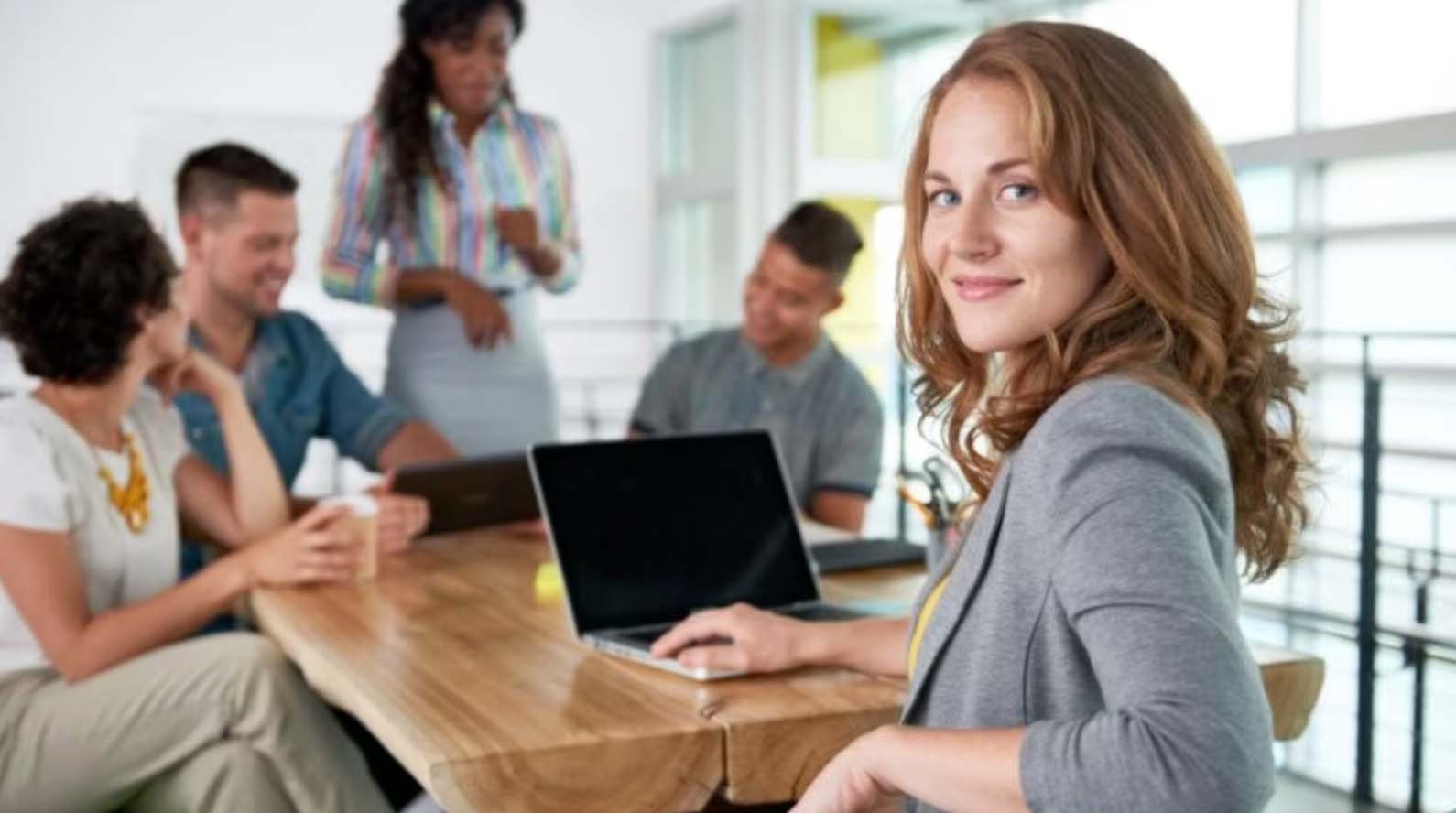 A woman at a laptop looking to camera while colleagues meet behind her