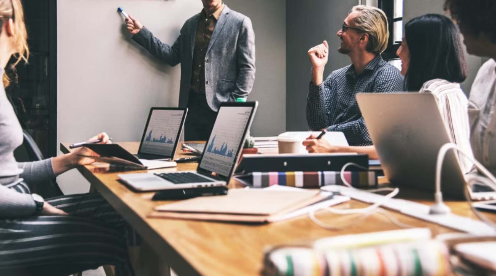 Team meeting around a table with laptops while a colleague presents at a whiteboard
