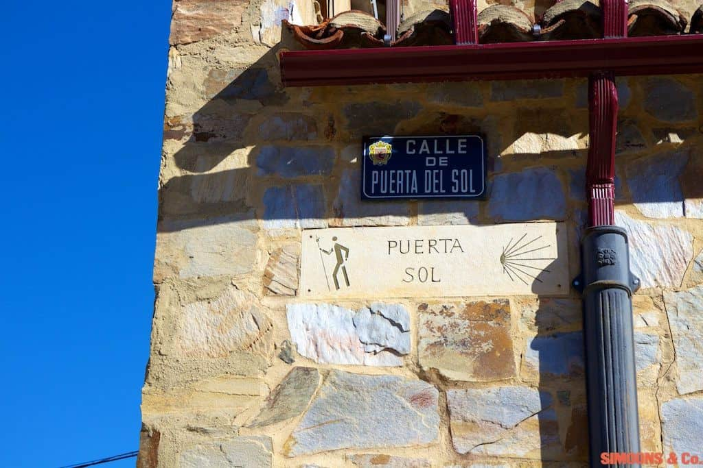 A Spanish street sign for Calle de Puerta del Sol beside a Camino de Santiago shell waymarker