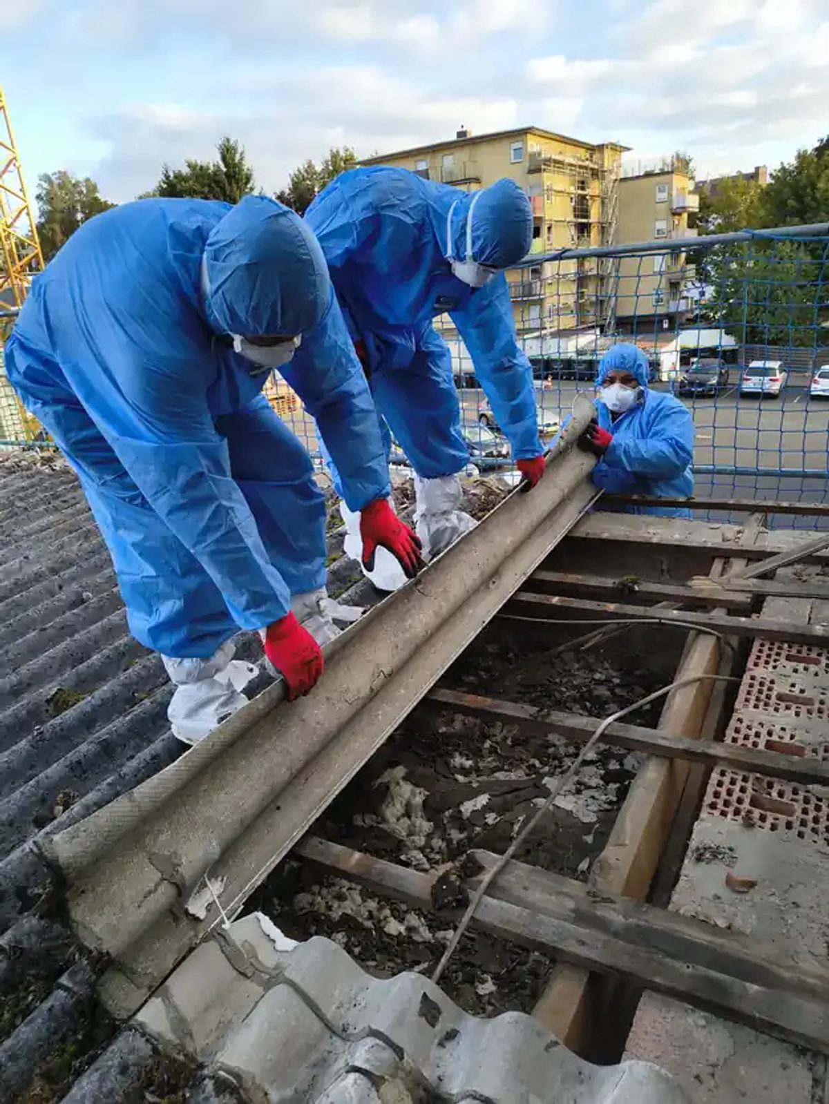 Drei Mitarbeiter in Schutzanzügen heben eine Asbestzement-Wellplatte von der freigelegten Dachlattung der Gewerbehalle in Kassel.
