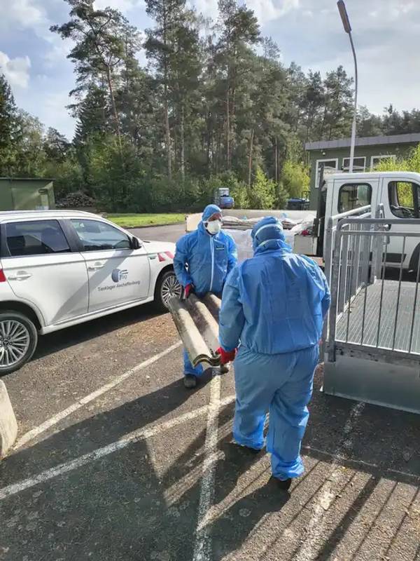 Zwei Mitarbeiter in Schutzanzügen tragen eine Asbestzement-Wellplatte zum Anhänger im NATO-Tanklager bei Aschaffenburg