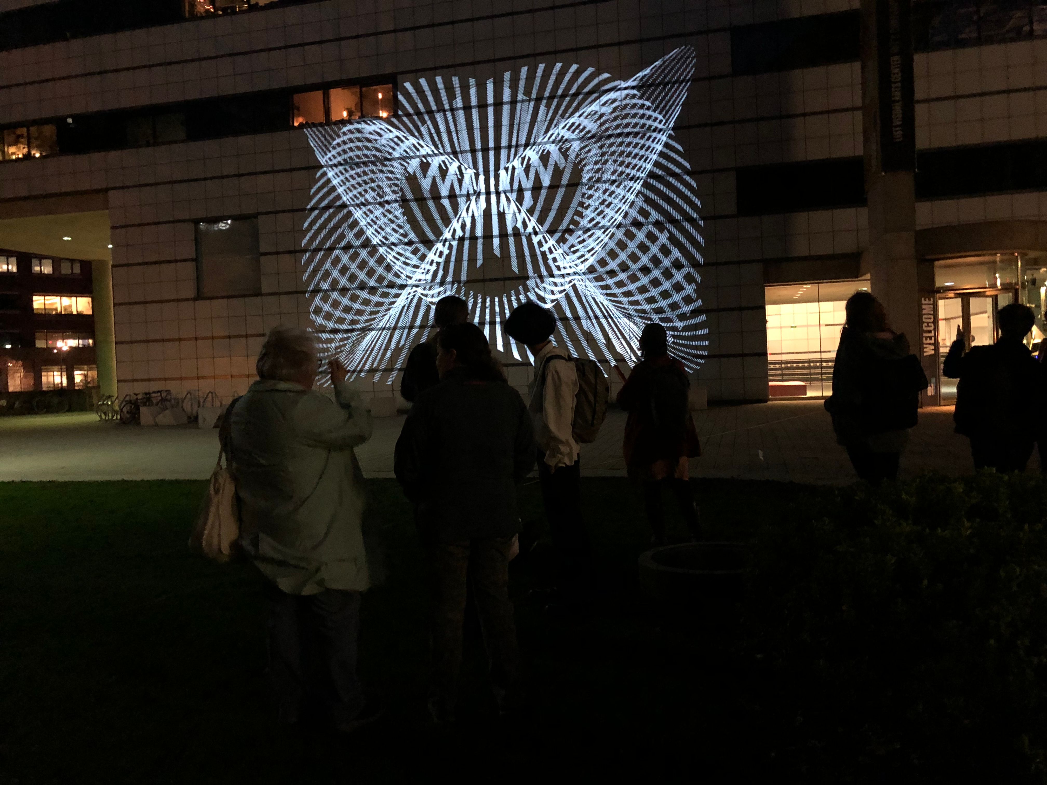 En gruppe mennesker står foran en lysprojeksjon på et hvitt bygg på kvelden