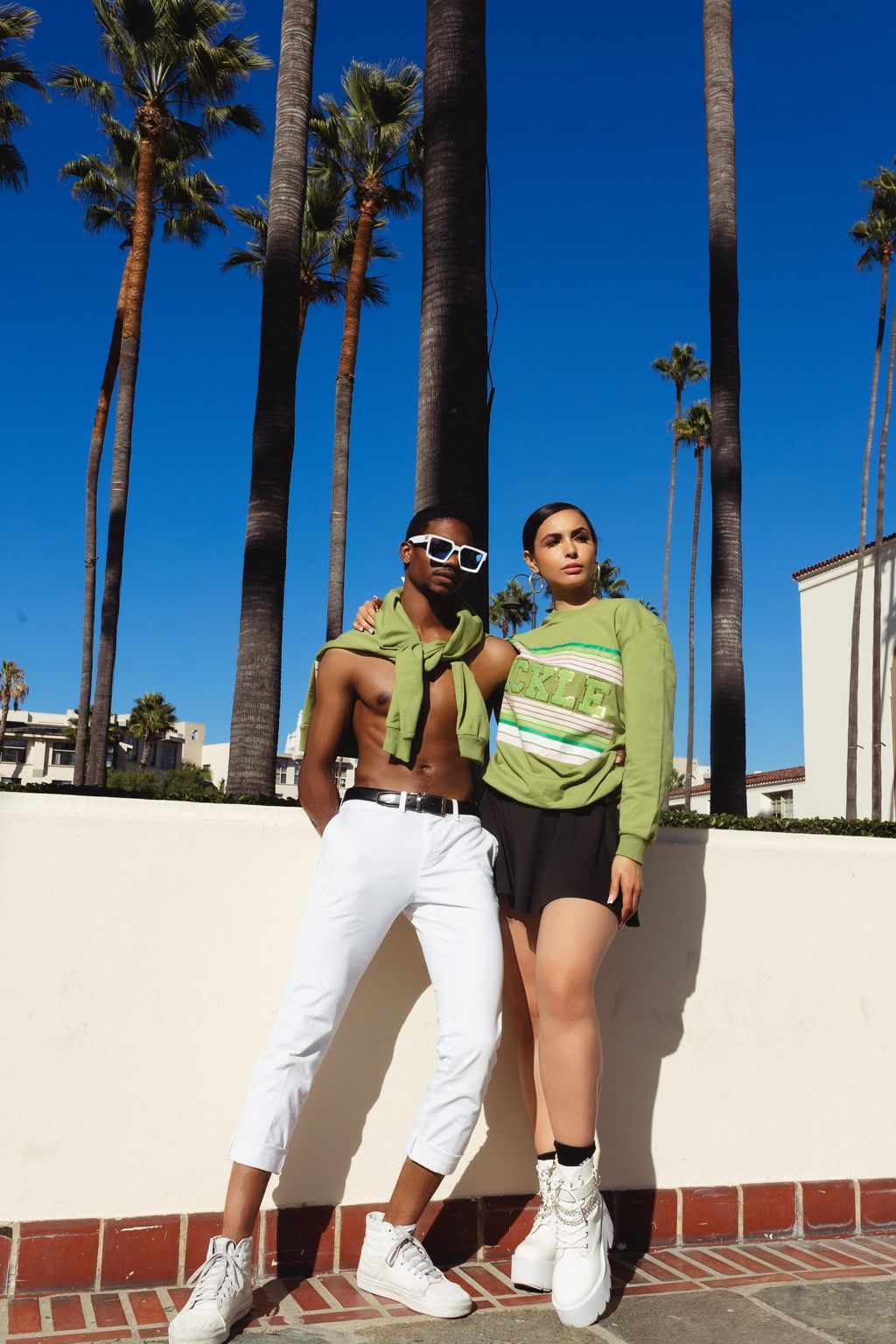 Two models in coordinated preppy knitwear standing together on a brick walkway lined with palms.
