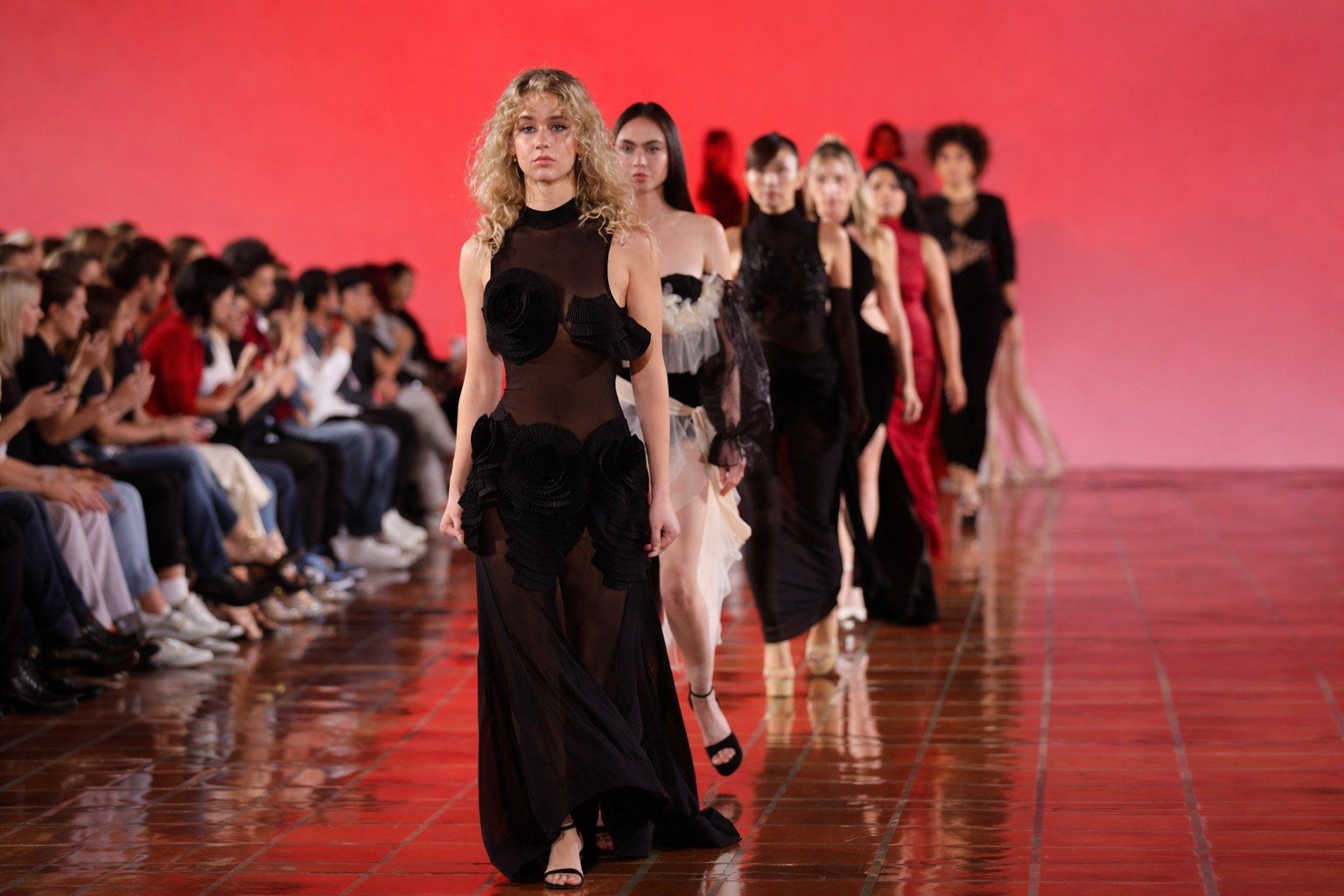 Finale walk at a fashion show: a line of models in black and red looks on a red-lit runway, with the front row of the audience seated along the edge.