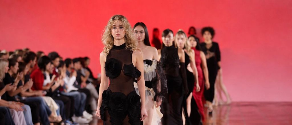 Finale walk at a fashion show: a line of models in black and red looks on a red-lit runway, with the front row of the audience seated along the edge.