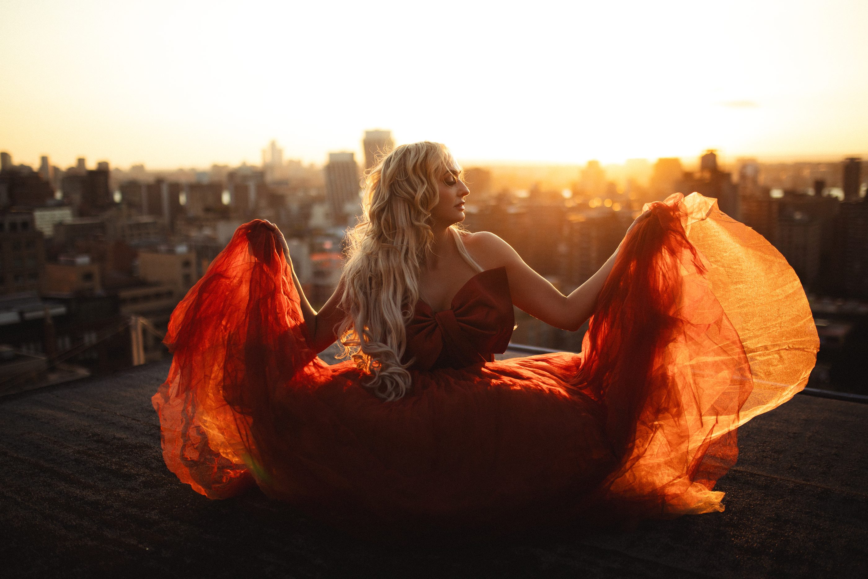Model with red fabric fanned out behind her, facing the sunset over Manhattan rooftops.