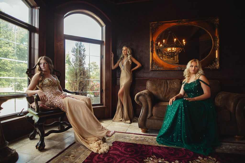 Three models in gowns gathered by tall arched windows in a dim manor interior.