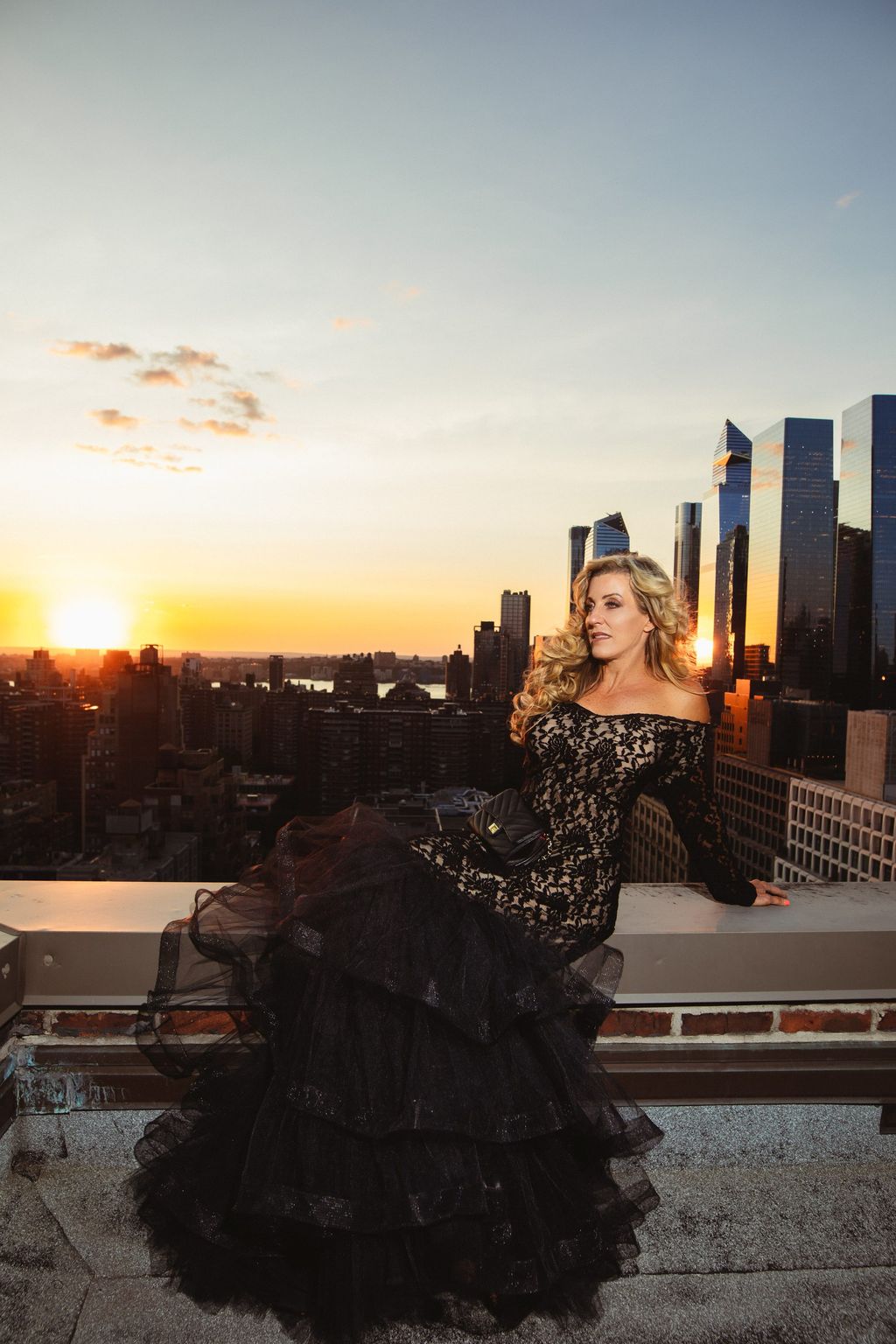 Model in a black lace gown seated on a rooftop ledge at dusk.