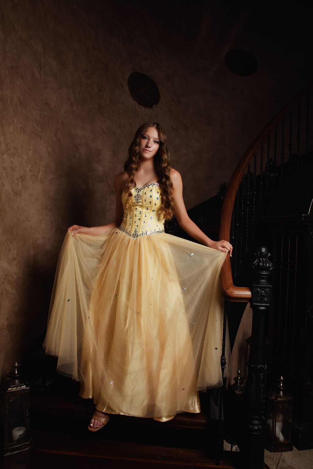 Model in a champagne tulle gown holding the skirt wide on a dark staircase.