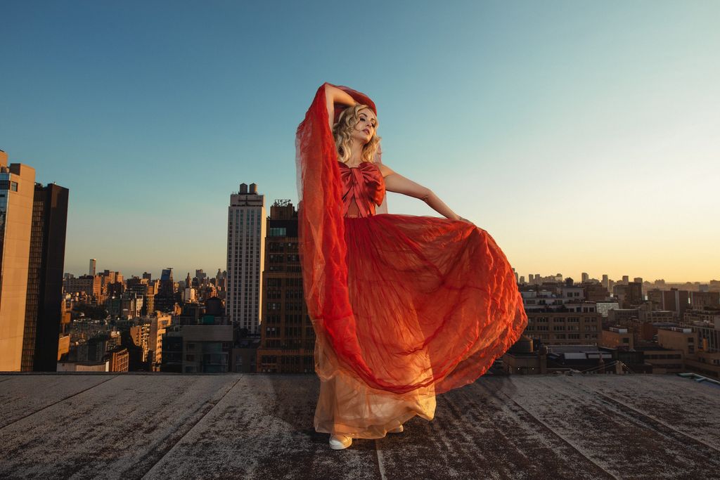 Model with flowing red-orange fabric billowing around her on a rooftop at golden hour.