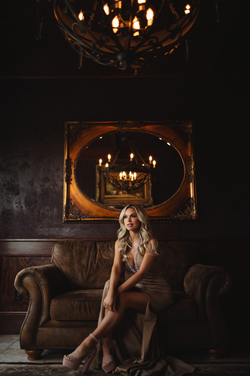 Model in a gold gown seated in an antique armchair beneath a chandelier in a dark wood-paneled room.