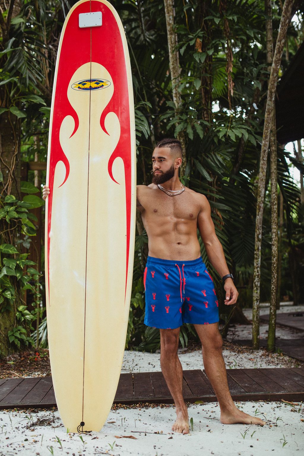 Man in blue swim trunks standing beside a flame-painted surfboard against the trees.
